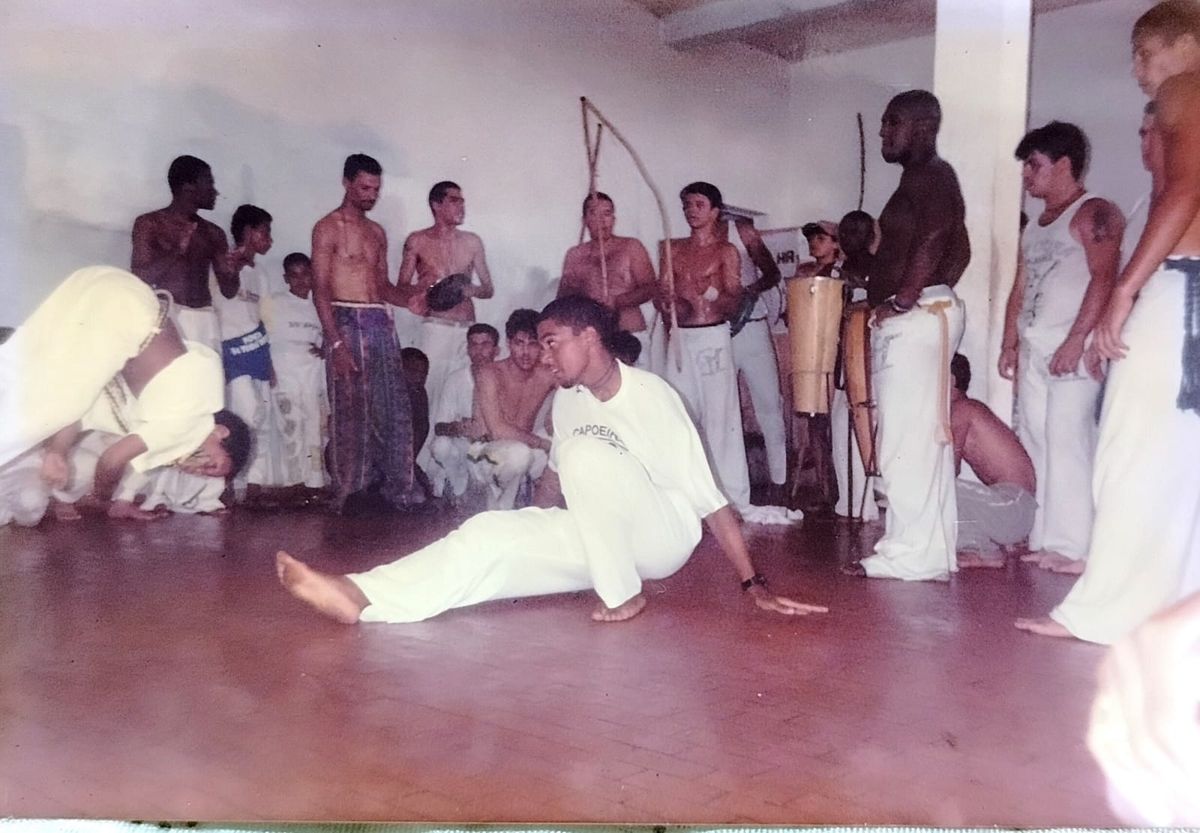 Roda de capoeira antiga, com capoeiristas de calça branca e bateria de berimbaus ao fundo